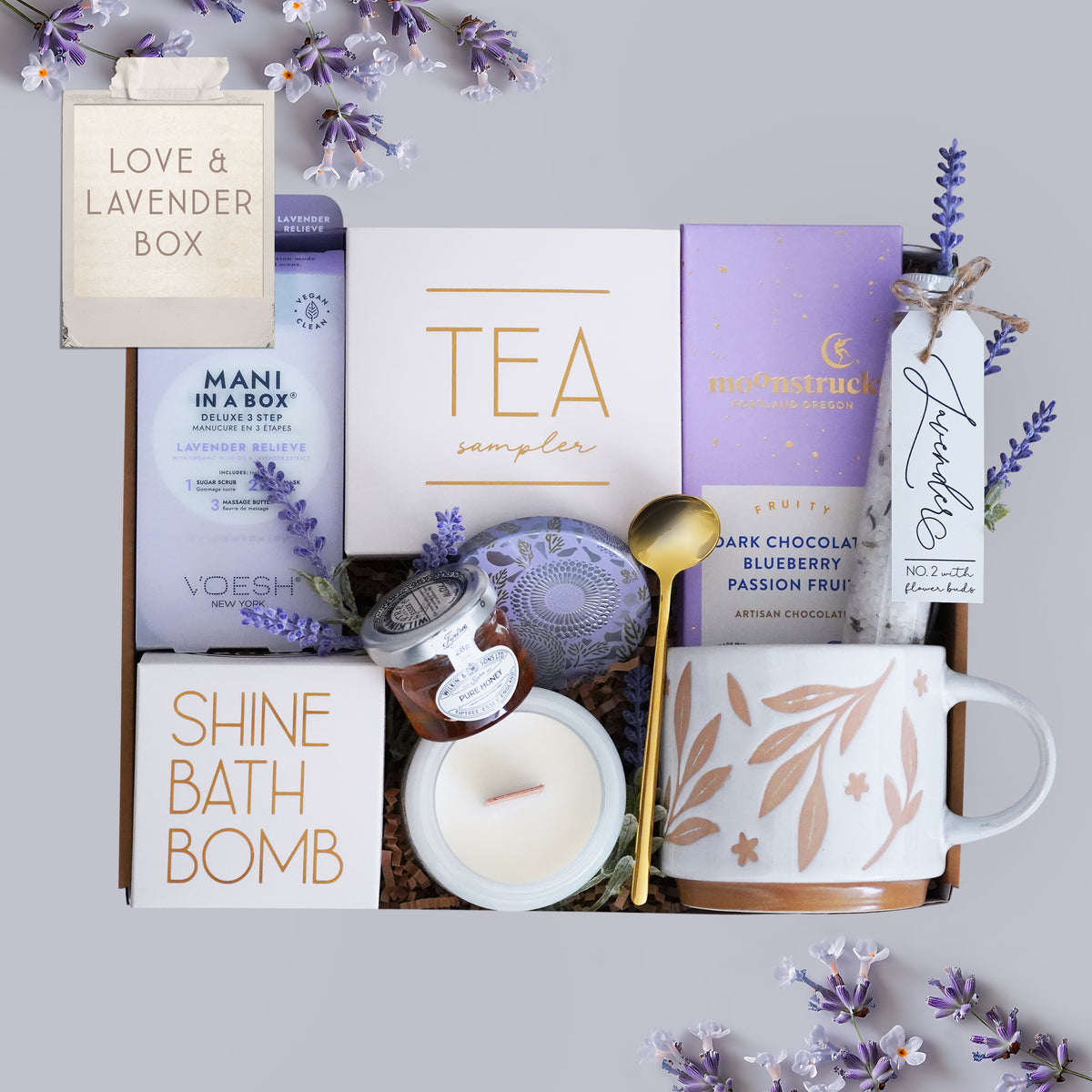 Gift box with tea, bath bomb, mug, and other items on a light gray background with lavender flowers.