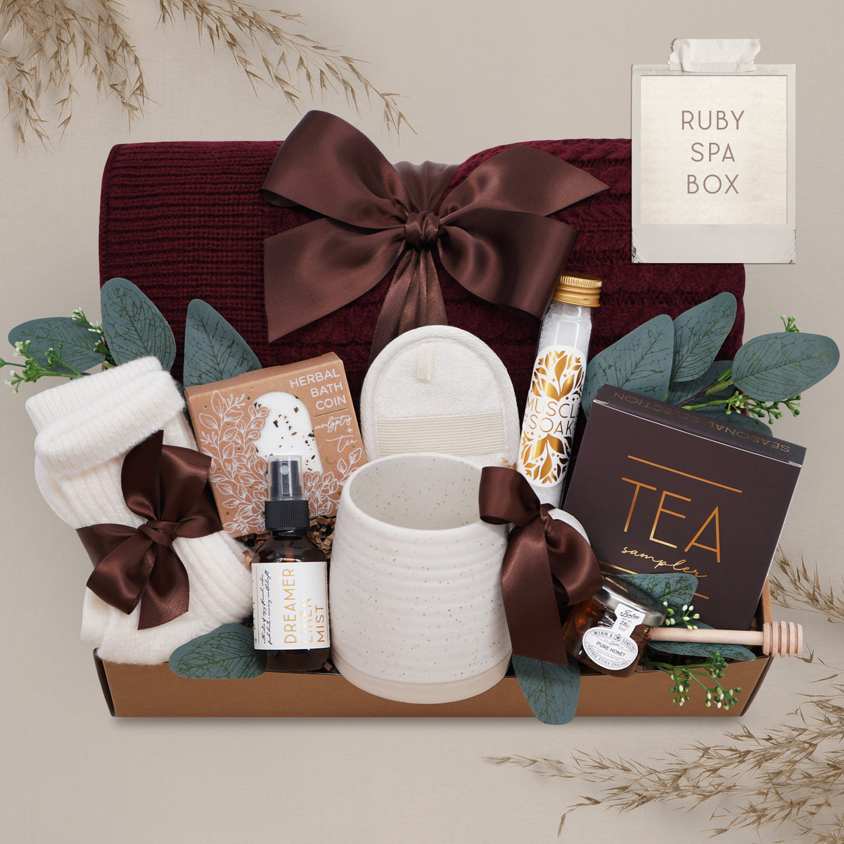 Wildflower Pop Ruby Spa Box with Burgundy blanket, bath salts, bath bomb, linen mist, cream socks, tea sampler, and mini honey and dipper. On tan grey background 