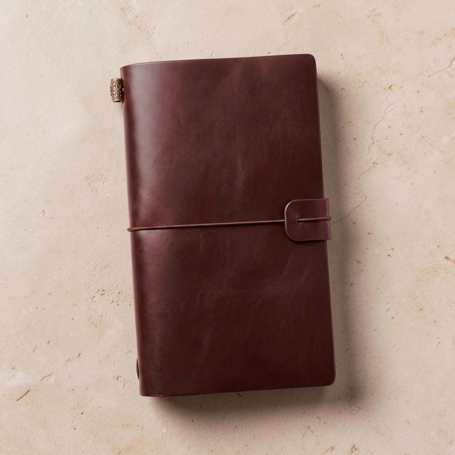 A dark brown faux leather travelers notebook journal with a wrap-around strap, photographed flat on a light beige textured surface.