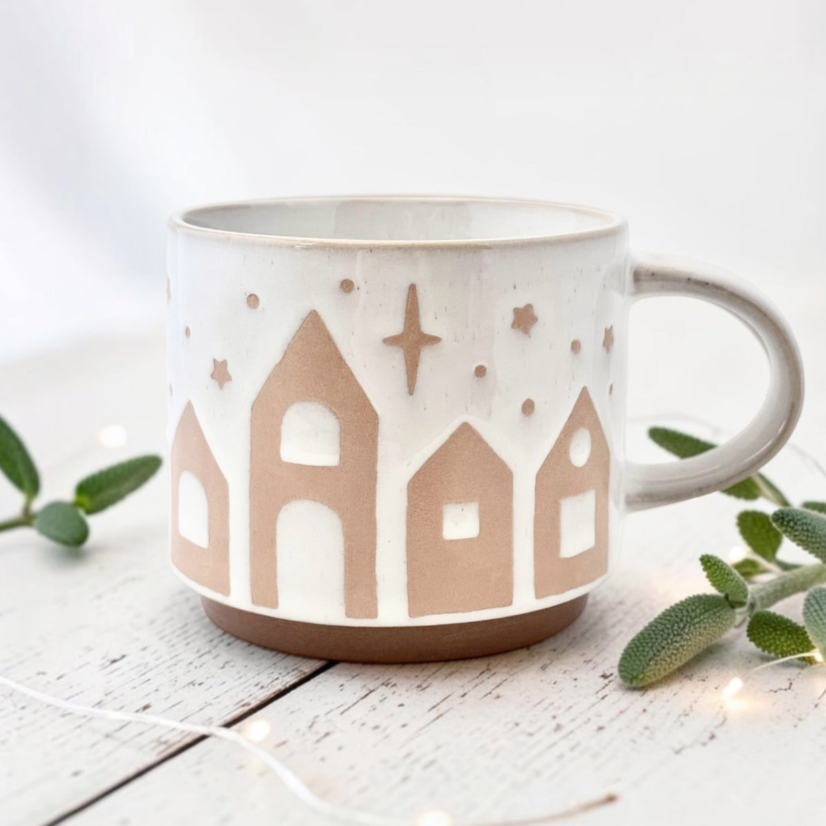 White ceramic mug with starry village design on a light wooden surface with greenery