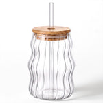 A clear ribbed glass tumbler with a wavy vertical shape, featuring a light natural wood lid and a clear plastic straw, shown on a white background.