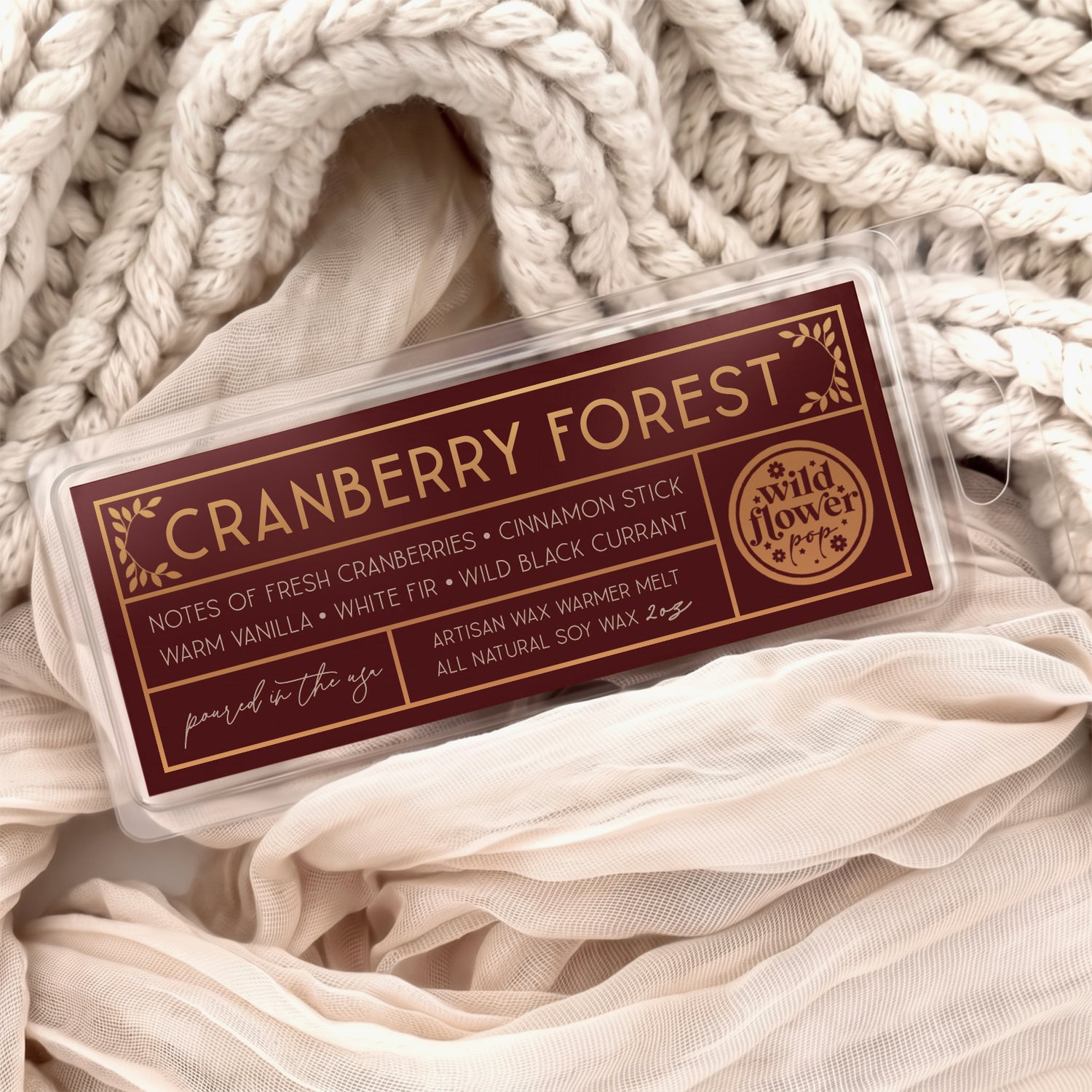 Cranberry Forest artisan wax melts, featuring a burgundy label with gold foil, resting on a cozy background of cream-colored softly draped fabric.