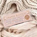 Orange Blossom artisan wax melts, featuring a pale pink label with gold foil, resting on a cozy background of cream-colored softly draped fabric.
