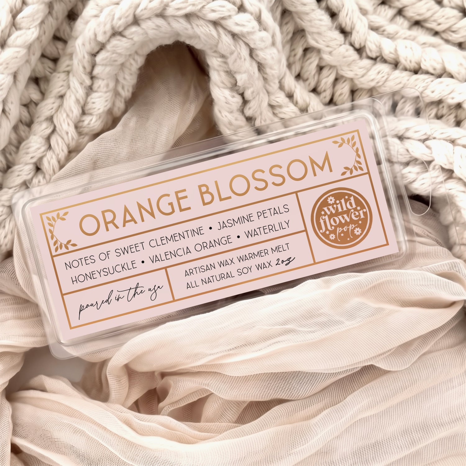 Orange Blossom artisan wax melts, featuring a pale pink label with gold foil, resting on a cozy background of cream-colored softly draped fabric.