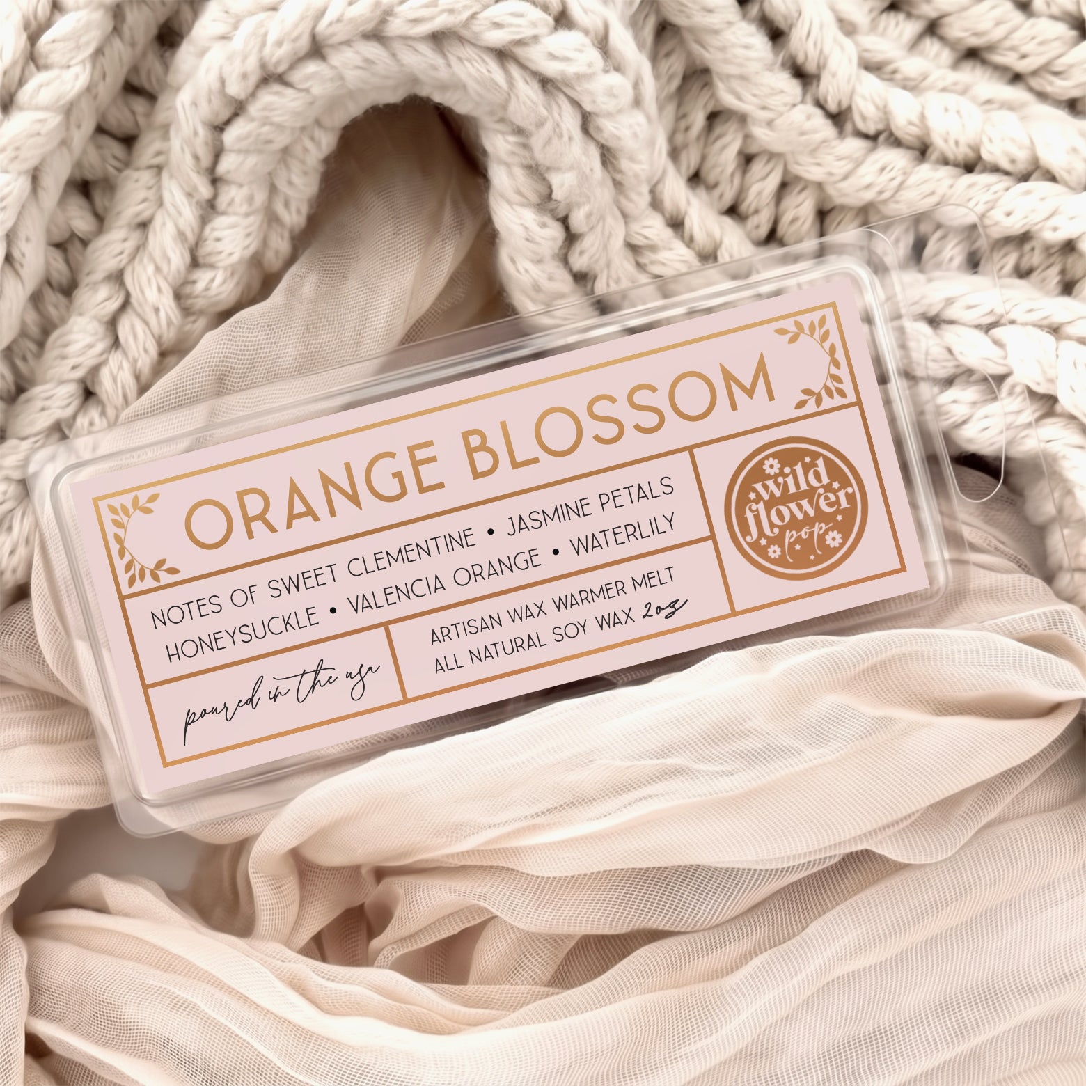 Orange Blossom artisan wax melts, featuring a pale pink label with gold foil, resting on a cozy background of cream-colored softly draped fabric.