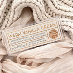 Warm Vanilla Cream artisan wax melts, featuring a cream label with gold foil, resting on a cozy background of cream-colored softly draped fabric.