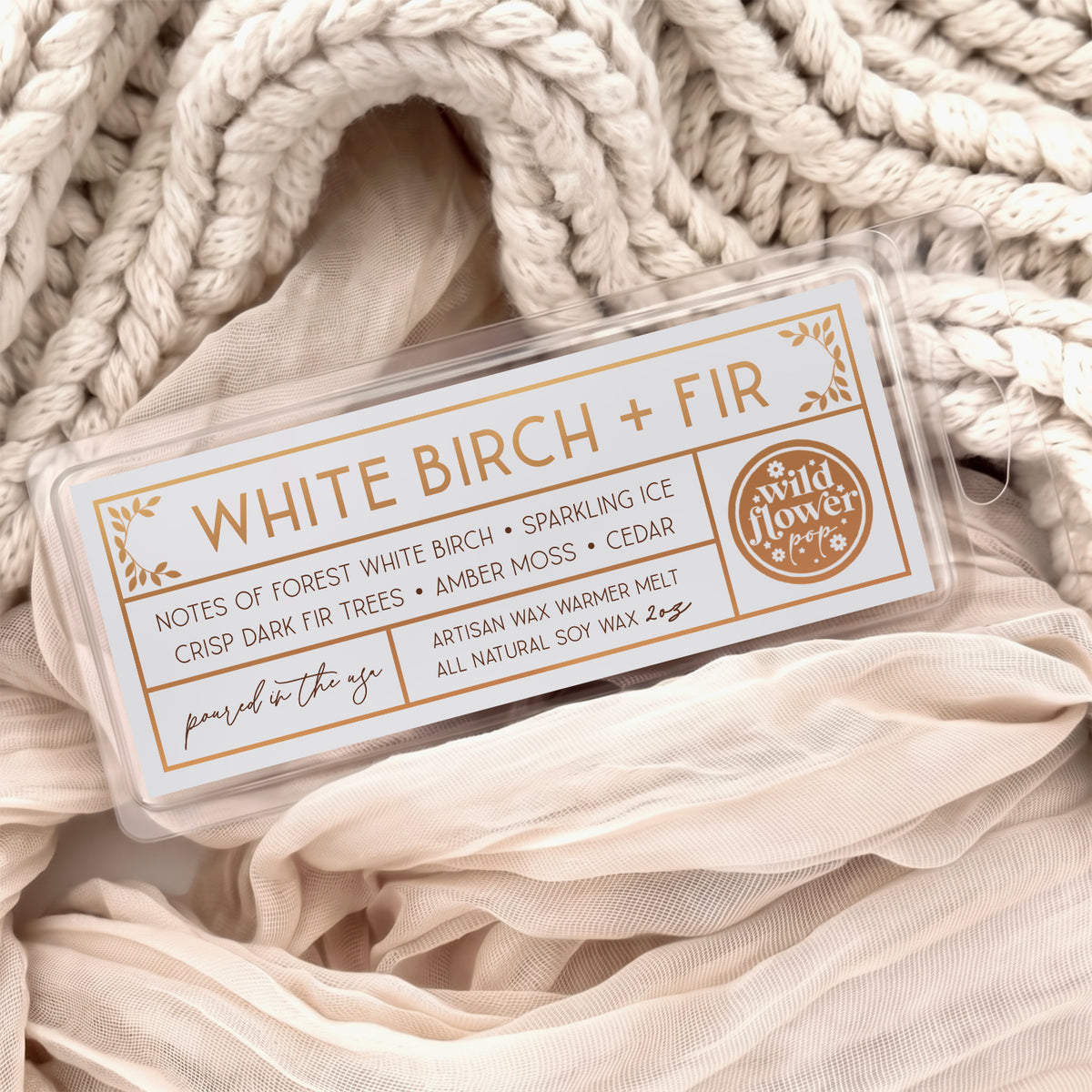 White Birch + Fir artisan wax melts, featuring a neutral gray label with gold foil, resting on a cozy background of cream-colored softly draped fabric.