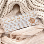 White Birch + Fir artisan wax melts, featuring a neutral gray label with gold foil, resting on a cozy background of cream-colored softly draped fabric.