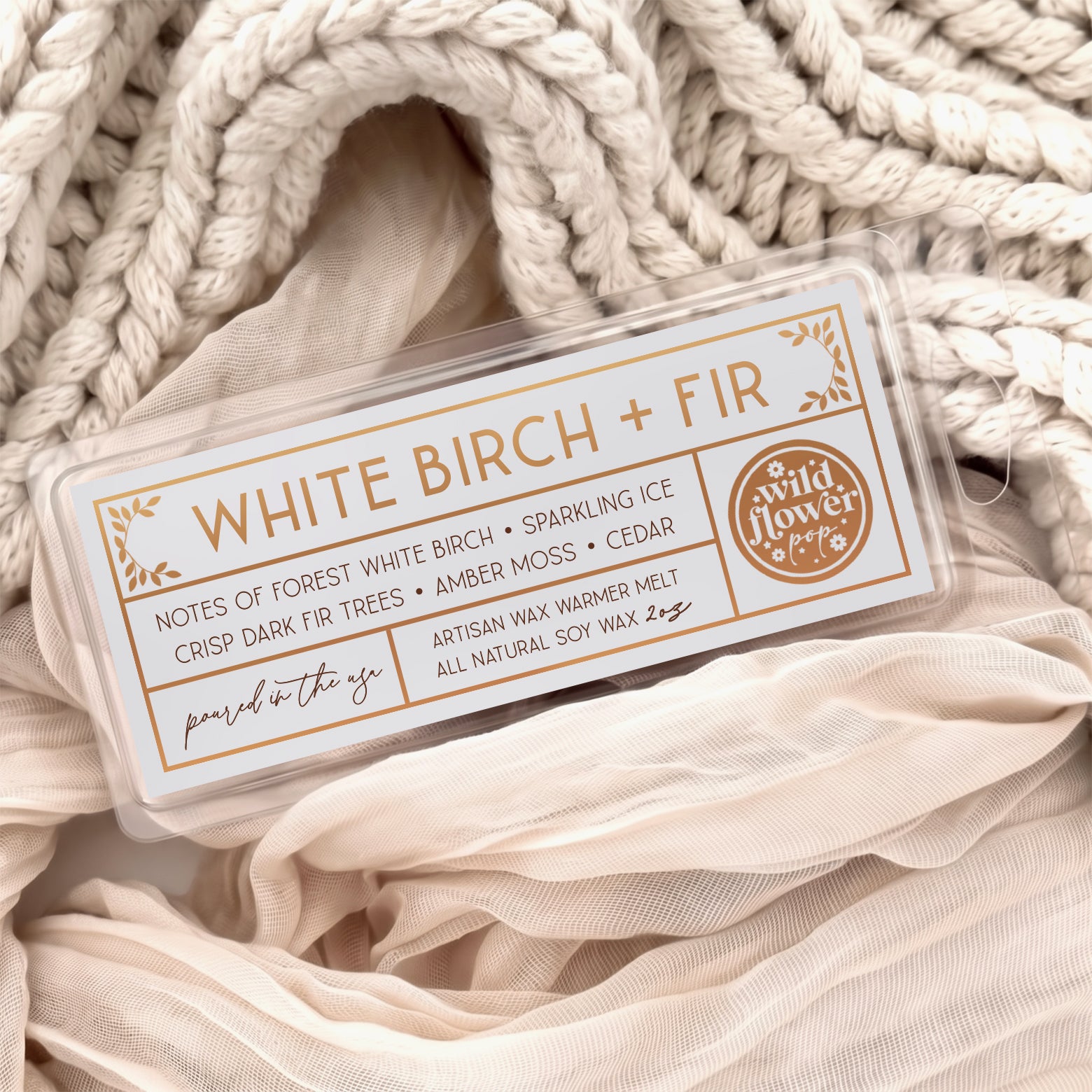 White Birch + Fir artisan wax melts, featuring a neutral gray label with gold foil, resting on a cozy background of cream-colored softly draped fabric.